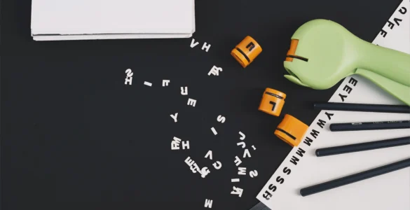 Uma mesa preta com letras e números espalhados, dois blocos laranja, um livro e lápis, sugerindo estudo.
