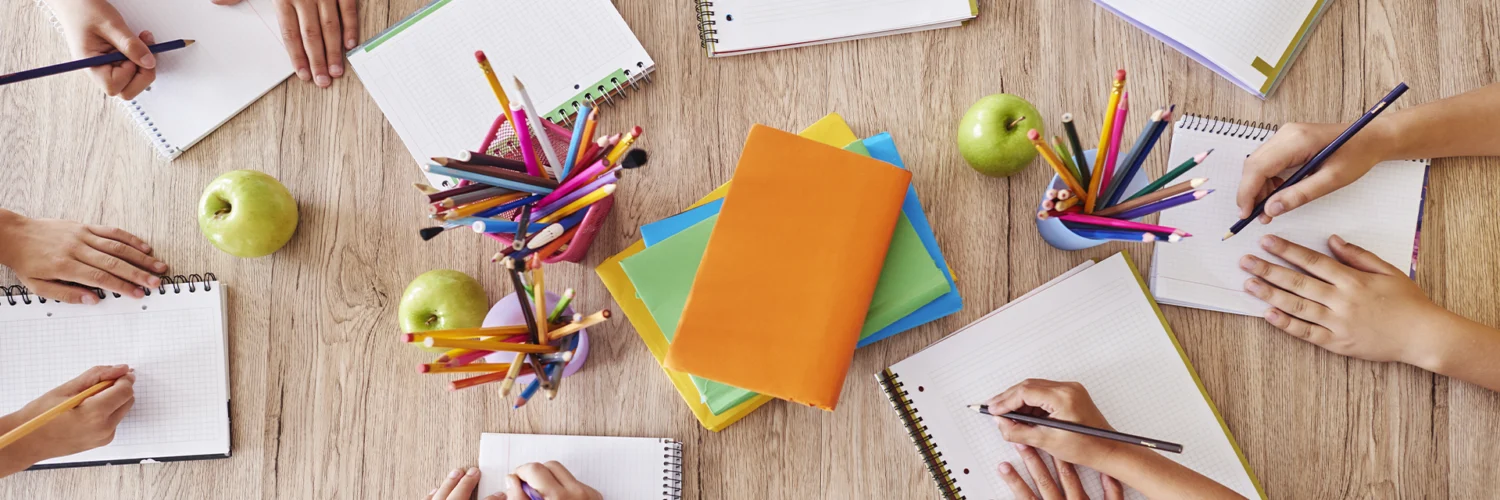 Crianças escrevendo em cadernos ao redor de uma mesa com lápis coloridos, livros e maçãs verdes.