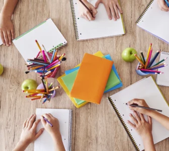 Crianças escrevendo em cadernos ao redor de uma mesa com lápis coloridos, livros e maçãs verdes.