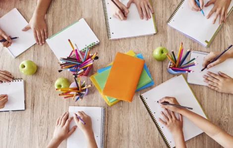 Crianças escrevendo em cadernos ao redor de uma mesa com lápis coloridos, livros e maçãs verdes.