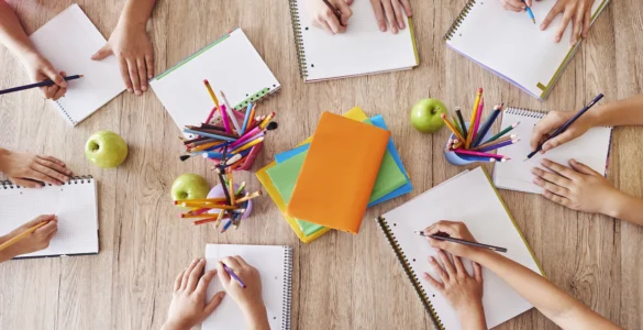 Crianças escrevendo em cadernos ao redor de uma mesa com lápis coloridos, livros e maçãs verdes.