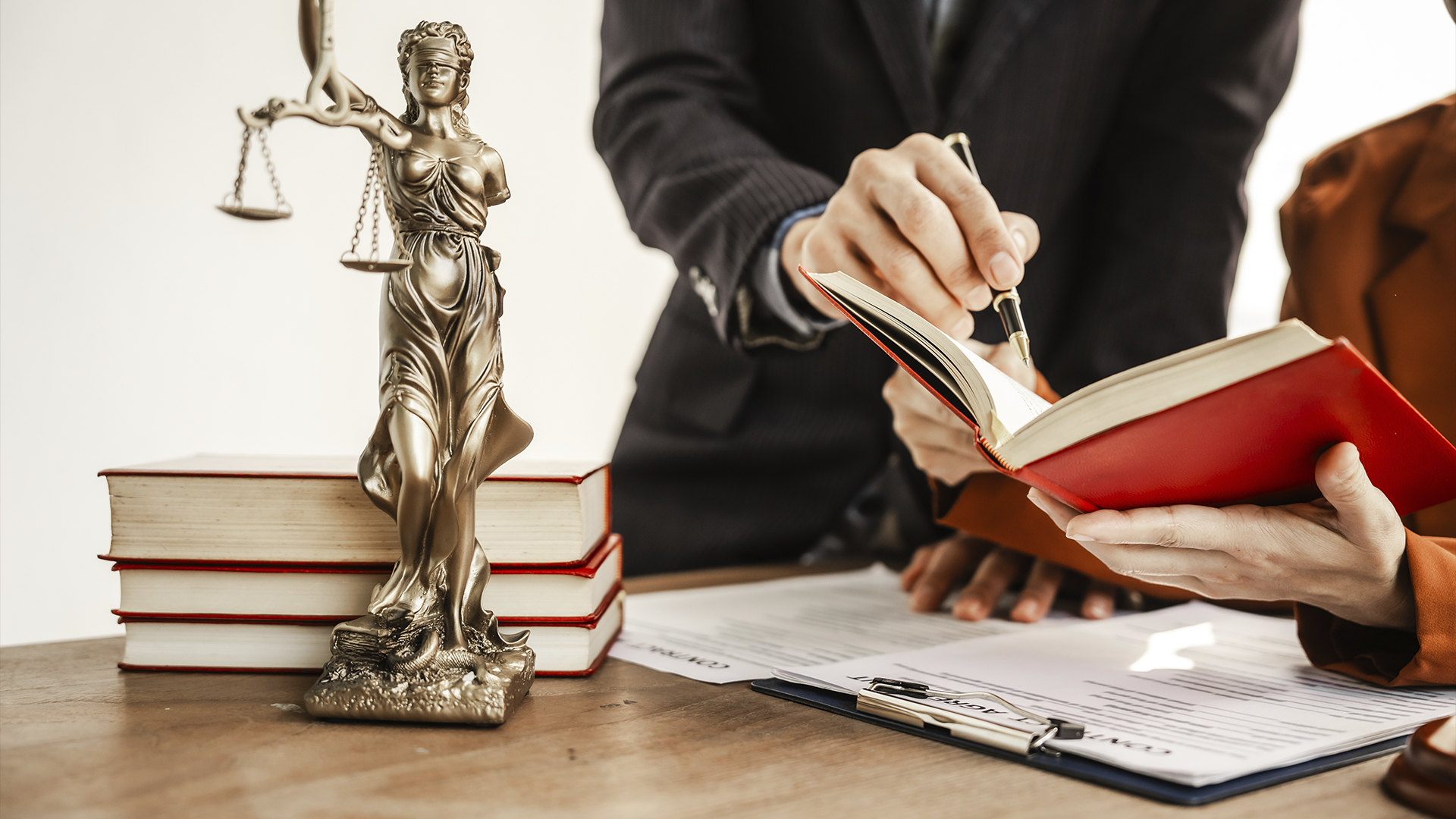 Estátua da justiça (Têmis) em metal dourado, livros e duas pessoas analisando um documento jurídico em uma mesa.
