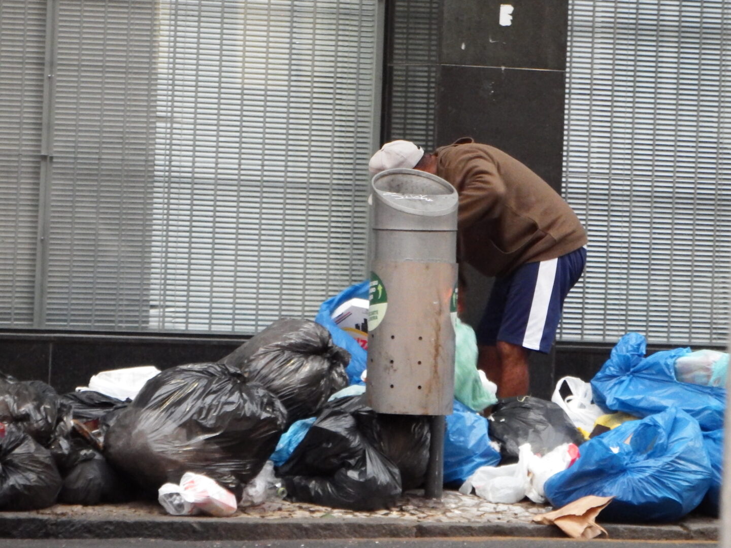 Homem em situação de rua revira lixo em calçada cercada por sacos de resíduos, em área urbana movimentada.