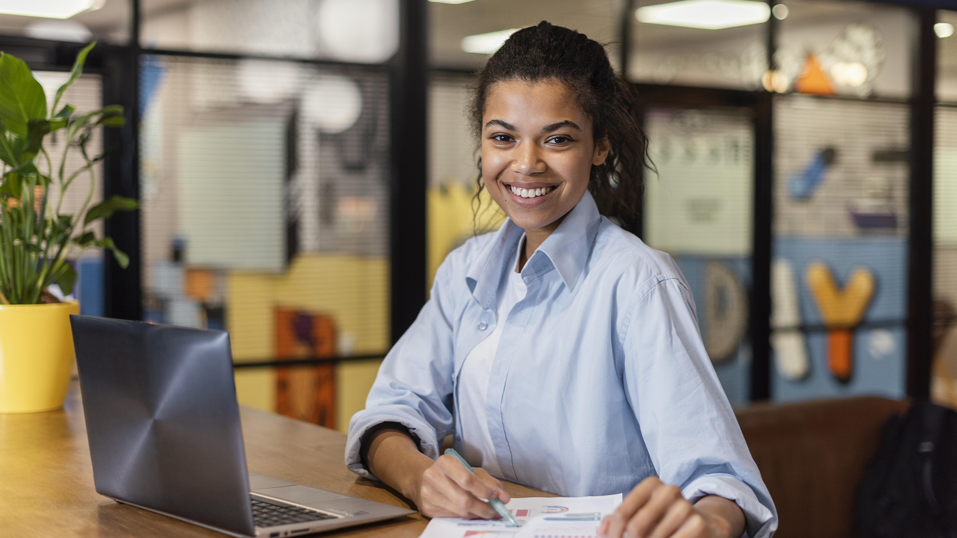 Uma mulher jovem e sorridente, de camisa azul, escrevendo em papéis com um laptop e planta ao lado.