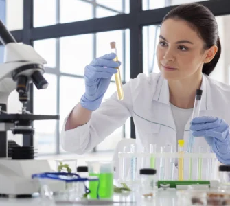 Pesquisadora analisa amostras em tubos de ensaio em laboratório, com luvas e microscópio ao lado.