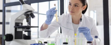 Pesquisadora analisa amostras em tubos de ensaio em laboratório, com luvas e microscópio ao lado.