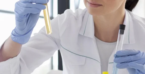 Pesquisadora analisa amostras em tubos de ensaio em laboratório, com luvas e microscópio ao lado.