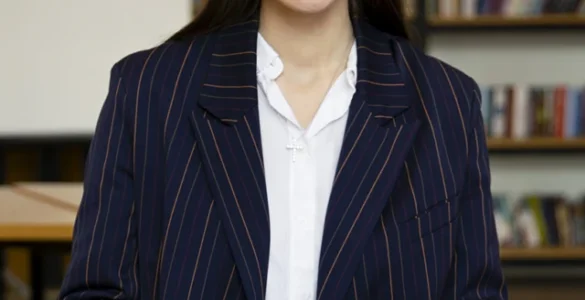 Uma mulher sorridente com óculos e blazer, sentada em uma mesa, escrevendo em papéis com um laptop ao lado.