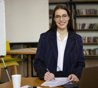 Uma mulher sorridente com óculos e blazer, sentada em uma mesa, escrevendo em papéis com um laptop ao lado.