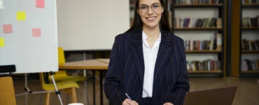 Uma mulher sorridente com óculos e blazer, sentada em uma mesa, escrevendo em papéis com um laptop ao lado.
