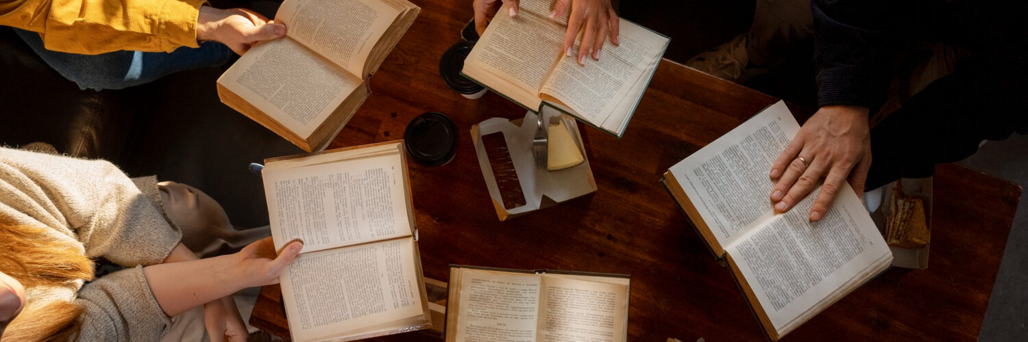 Grupo de pessoas sentado em torno de mesa de madeira, lendo livros abertos e tomando café.