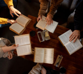 Grupo de pessoas sentado em torno de mesa de madeira, lendo livros abertos e tomando café.
