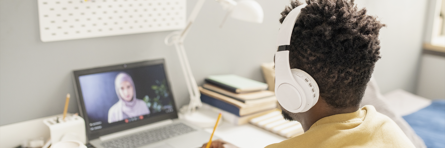 Pessoa com fones de ouvido assiste a aula online em laptop, escrevendo em caderno sobre a mesa organizada.