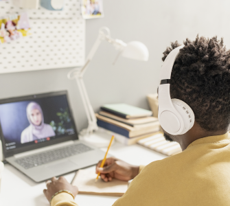 Pessoa com fones de ouvido assiste a aula online em laptop, escrevendo em caderno sobre a mesa organizada.