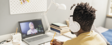 Pessoa com fones de ouvido assiste a aula online em laptop, escrevendo em caderno sobre a mesa organizada.