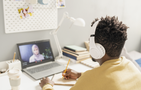 Pessoa com fones de ouvido assiste a aula online em laptop, escrevendo em caderno sobre a mesa organizada.