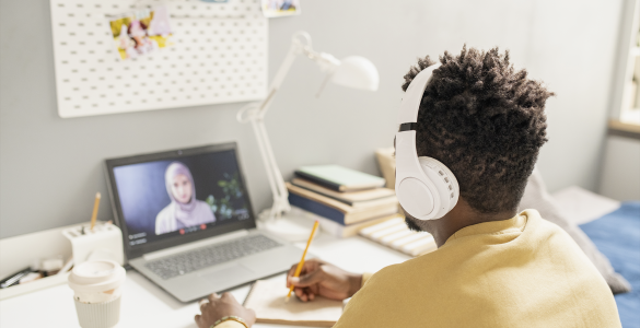 Pessoa com fones de ouvido assiste a aula online em laptop, escrevendo em caderno sobre a mesa organizada.