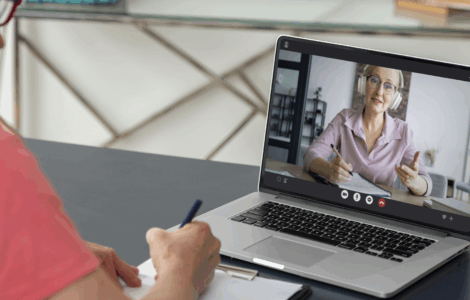 Pessoa participa de aula online pelo notebook, enquanto faz anotações em um caderno durante a videochamada.
