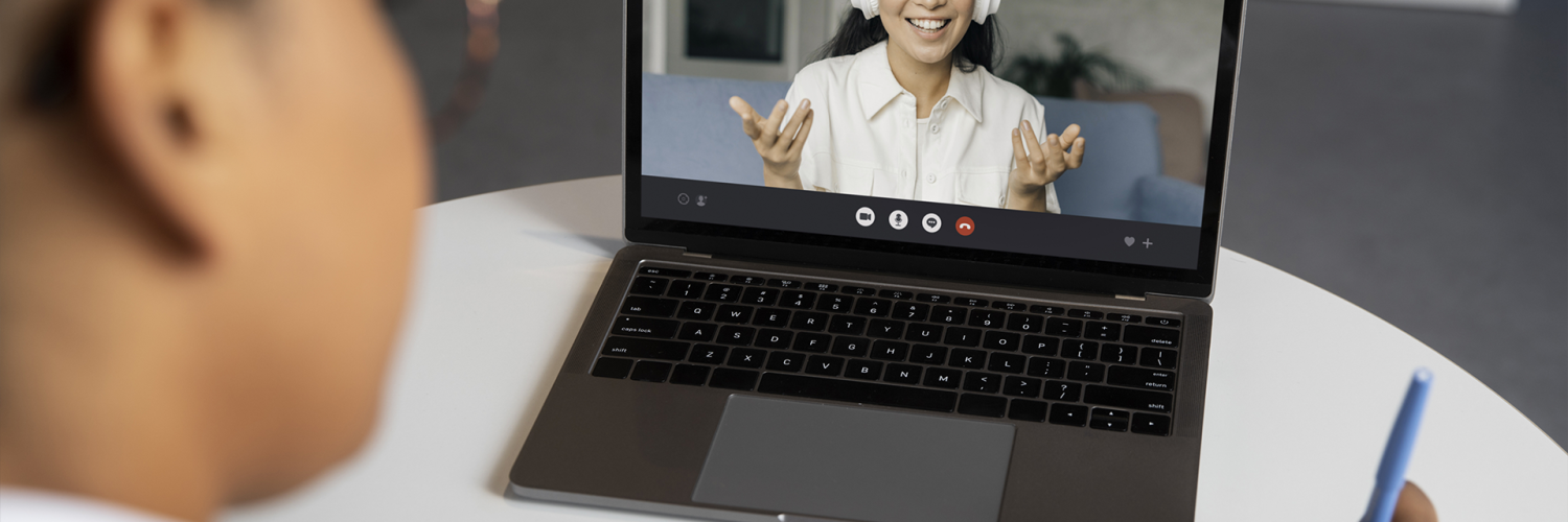 Pessoa participa de videochamada em laptop enquanto anota em caderno, interagindo com outra pessoa na tela.