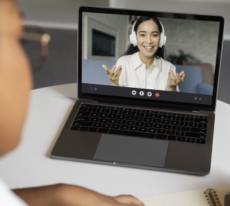 Pessoa participa de videochamada em laptop enquanto anota em caderno, interagindo com outra pessoa na tela.