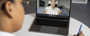 Pessoa participa de videochamada em laptop enquanto anota em caderno, interagindo com outra pessoa na tela.