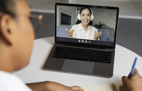 Pessoa participa de videochamada em laptop enquanto anota em caderno, interagindo com outra pessoa na tela.