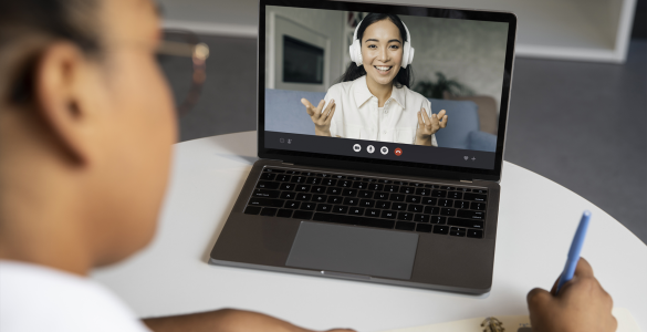 Pessoa participa de videochamada em laptop enquanto anota em caderno, interagindo com outra pessoa na tela.