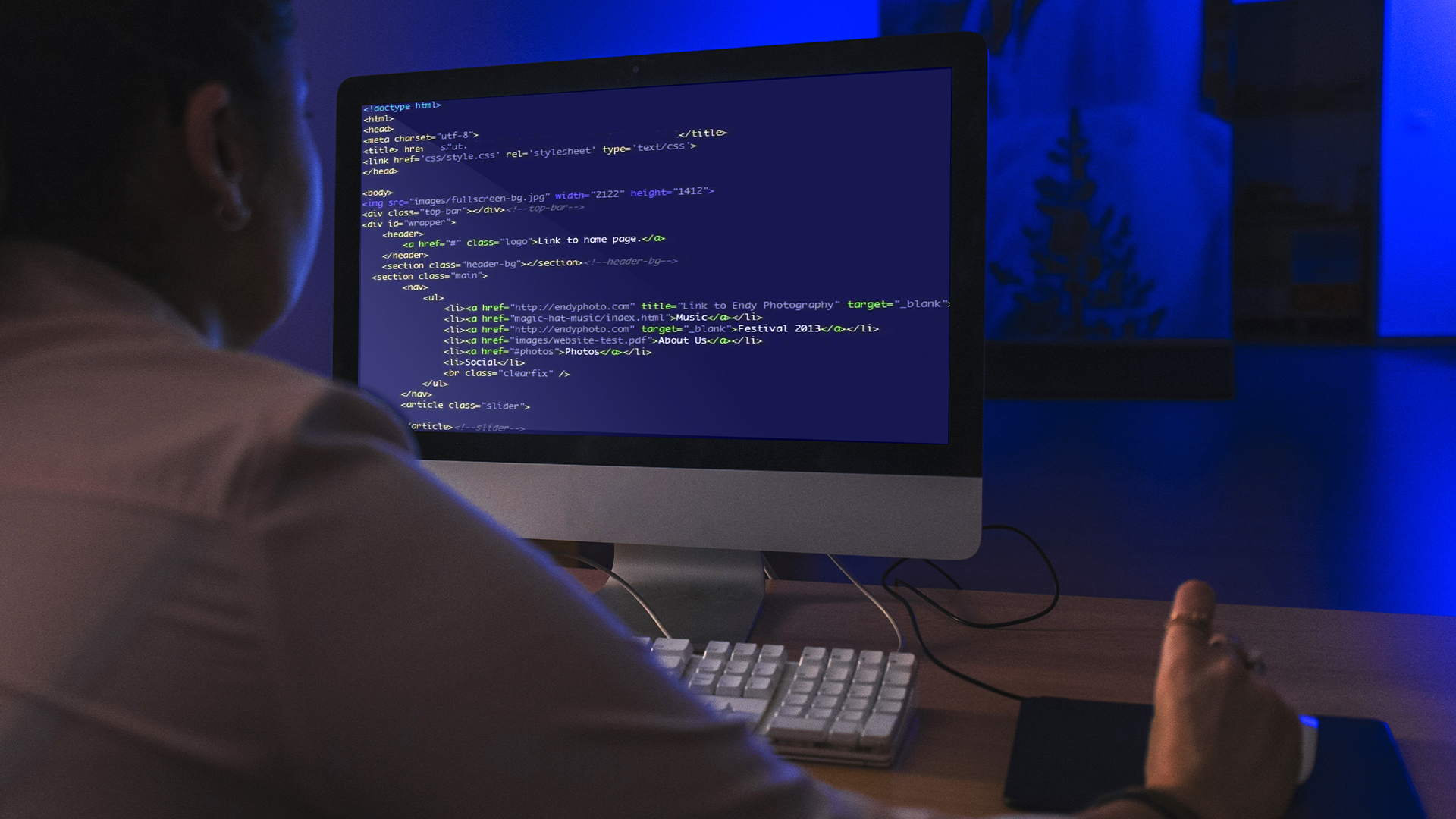 Pessoa programando em desktop, com códigos em HTML e CSS exibidos na tela em ambiente iluminado de azul.