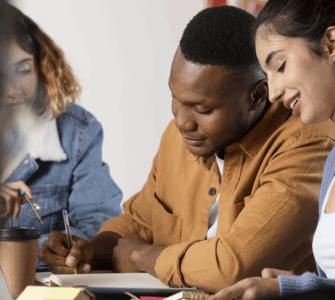 Grupo de jovens estudando juntos em mesa, trocando ideias e escrevendo em cadernos com atenção e colaboração.