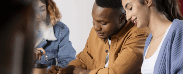Grupo de jovens estudando juntos em mesa, trocando ideias e escrevendo em cadernos com atenção e colaboração.