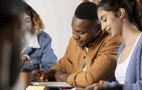 Grupo de jovens estudando juntos em mesa, trocando ideias e escrevendo em cadernos com atenção e colaboração.