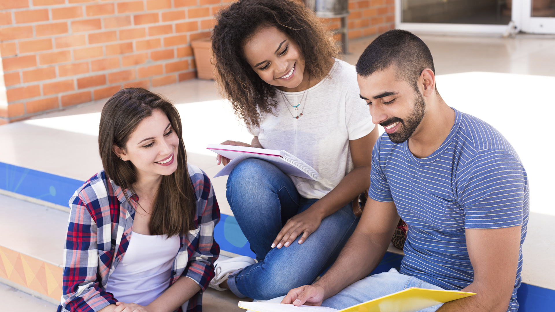 Três jovens sentados em escada ao ar livre, sorrindo e estudando juntos com cadernos e anotações.