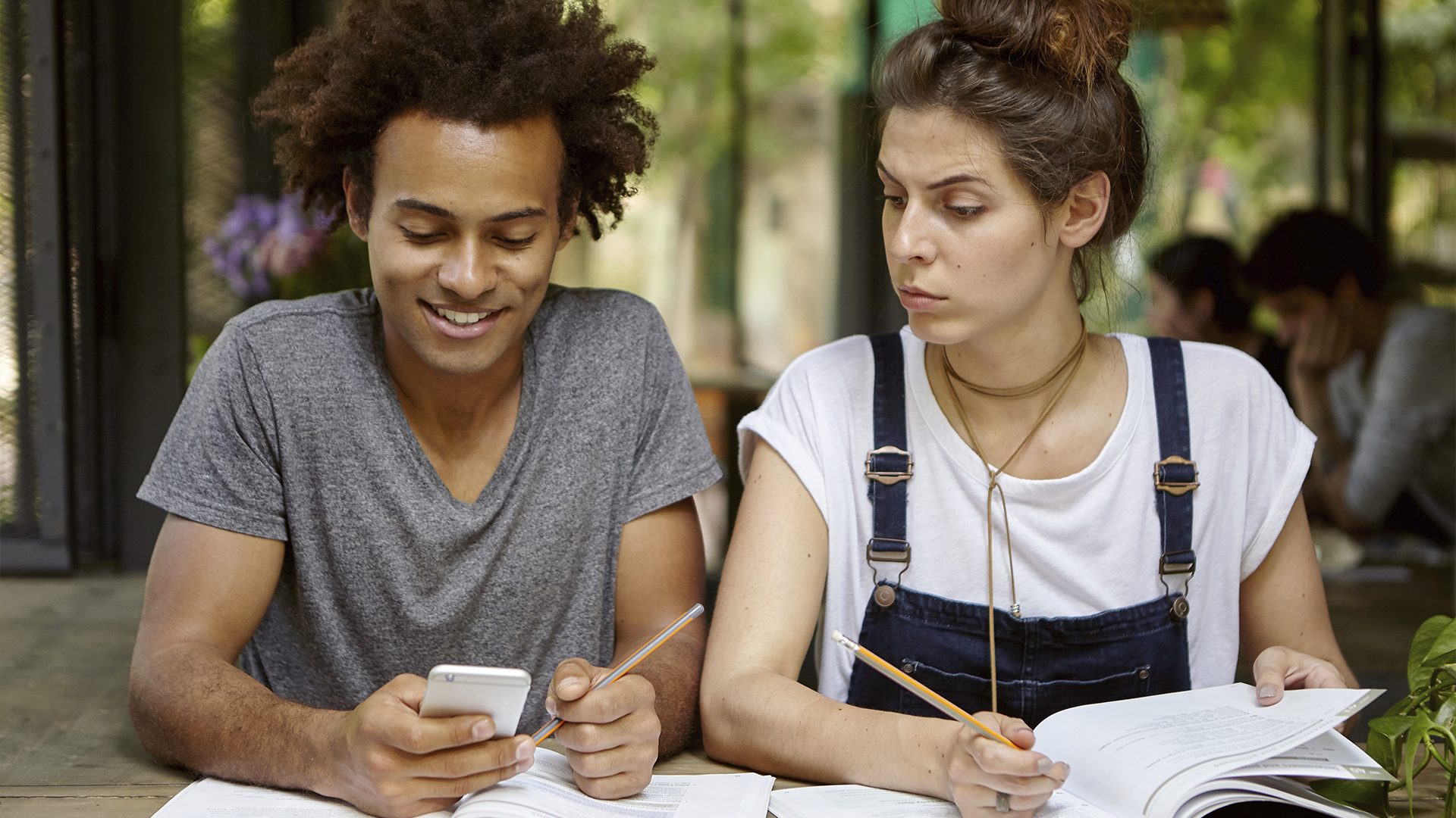 Dois jovens estudam juntos, um sorrindo enquanto usa o celular e o outro olhando desconfiado com livro aberto.