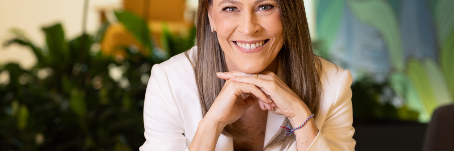 Ana Claudia Plihal sorridente de blazer branco em ambiente corporativo, transmitindo profissionalismo e confiança.