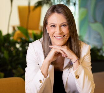 Ana Claudia Plihal sorridente de blazer branco em ambiente corporativo, transmitindo profissionalismo e confiança.