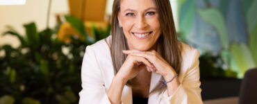 Ana Claudia Plihal sorridente de blazer branco em ambiente corporativo, transmitindo profissionalismo e confiança.