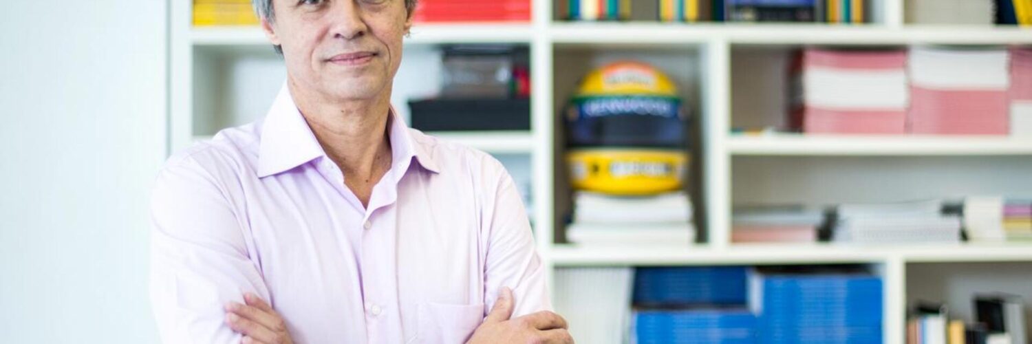 Foto de Mozart Neves Ramos, entrevistado, na frente de uma estante cheia de livros coloridos.