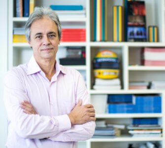 Foto de Mozart Neves Ramos, entrevistado, na frente de uma estante cheia de livros coloridos.