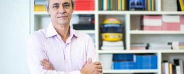 Foto de Mozart Neves Ramos, entrevistado, na frente de uma estante cheia de livros coloridos.