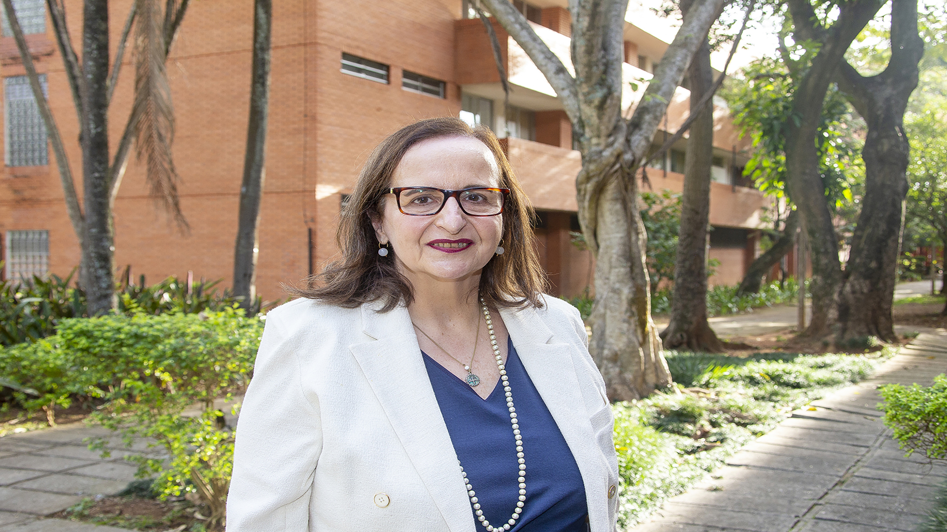 Foto da entrevistada Carlota Boto, em um campus da USP. Ao fundo, um prédio alaranjado em meio a árvores.