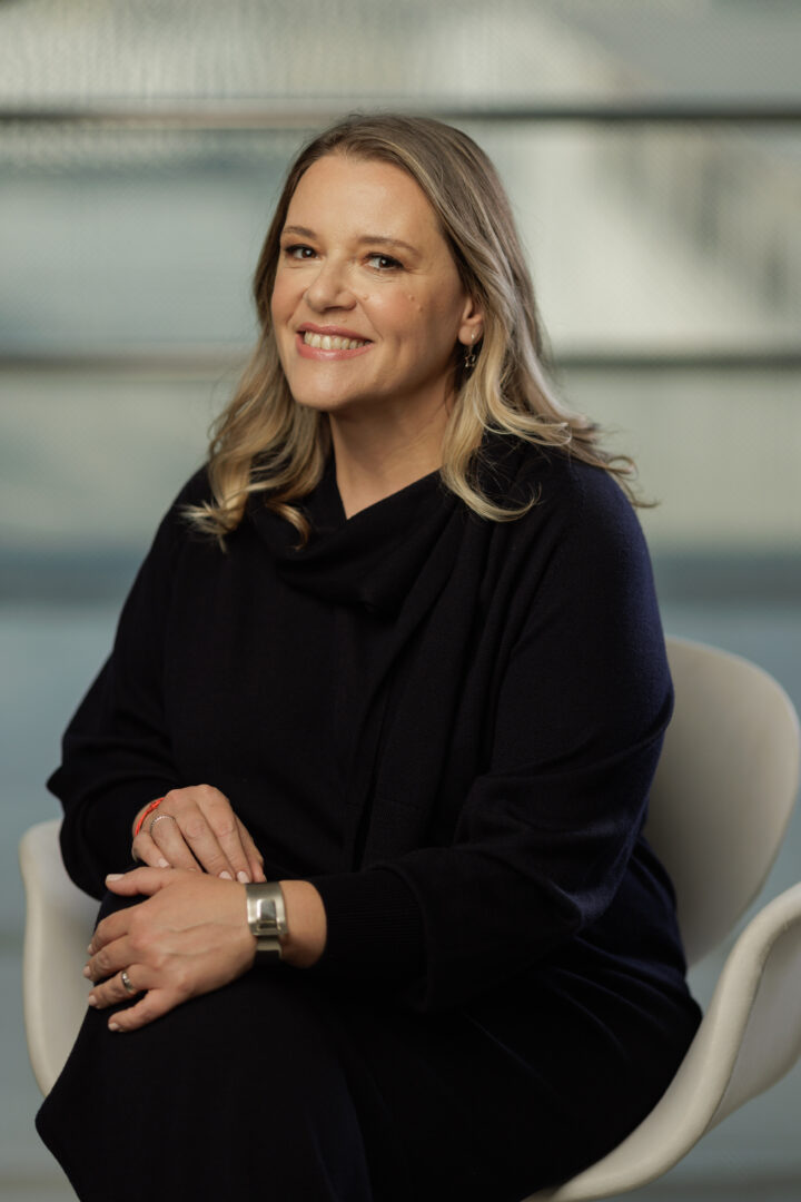 Vivian Broge sorridente com vestido preto sentada em cadeira moderna, transmitindo profissionalismo e confiança.