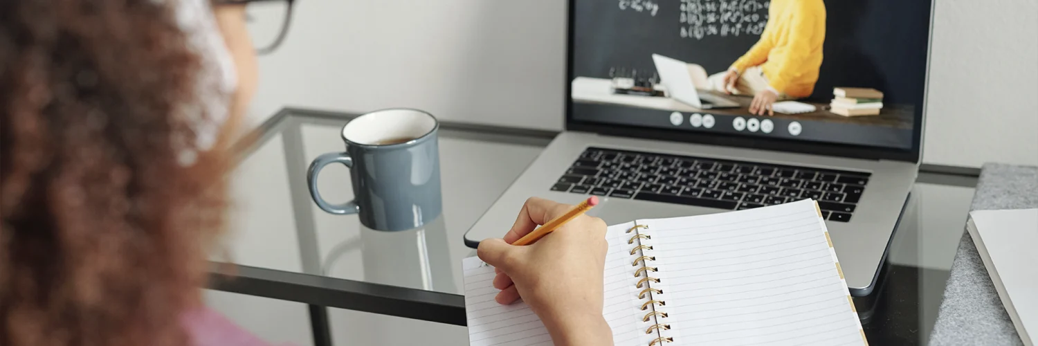 Pessoa assiste aula online em laptop e faz anotações em caderno, com caneca de café ao lado.