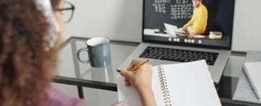 Pessoa assiste aula online em laptop e faz anotações em caderno, com caneca de café ao lado.
