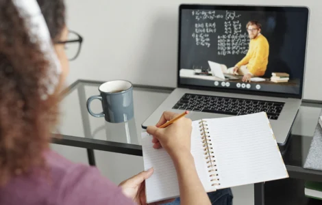 Pessoa assiste aula online em laptop e faz anotações em caderno, com caneca de café ao lado.