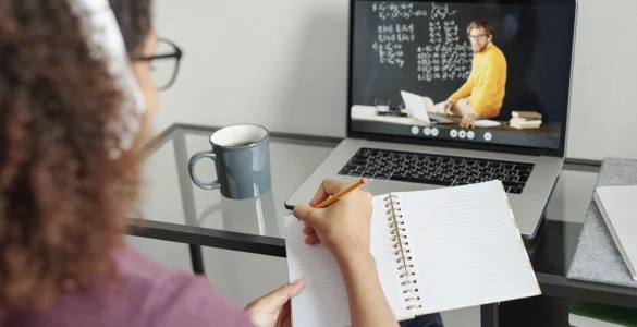 Pessoa assiste aula online em laptop e faz anotações em caderno, com caneca de café ao lado.