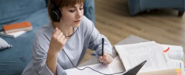 Estudante com fones de ouvido anota em caderno enquanto estuda online com livros abertos e notebook à frente.