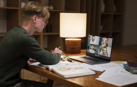 Estudante em videoconferência no notebook, estudando com livros e anotações sobre ensino a distância.