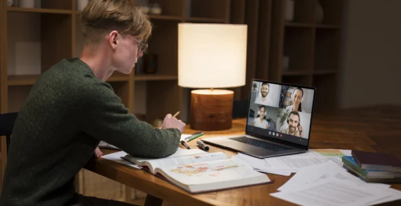 Estudante em videoconferência no notebook, estudando com livros e anotações sobre ensino a distância.