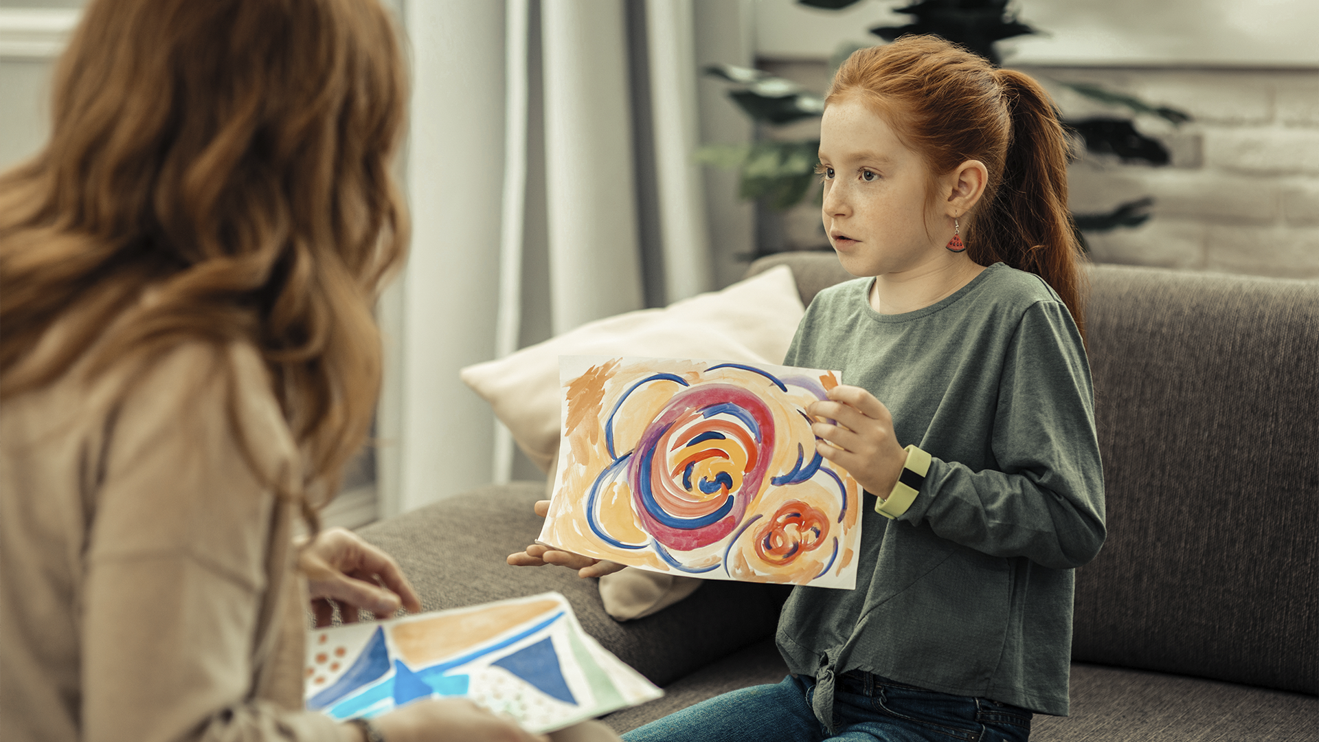 Uma menina ruiva segura um desenho colorido enquanto conversa com um adulto em um sofá, de frente para a câmera.