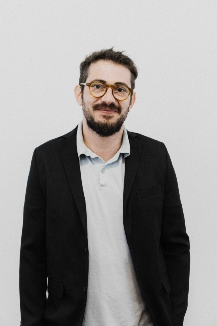 Foto do entrevistado, Prof. Me. Joaquim Ferreira Mendes Neto, em um fundo branco, usando roupas sociais formais e sorrindo.